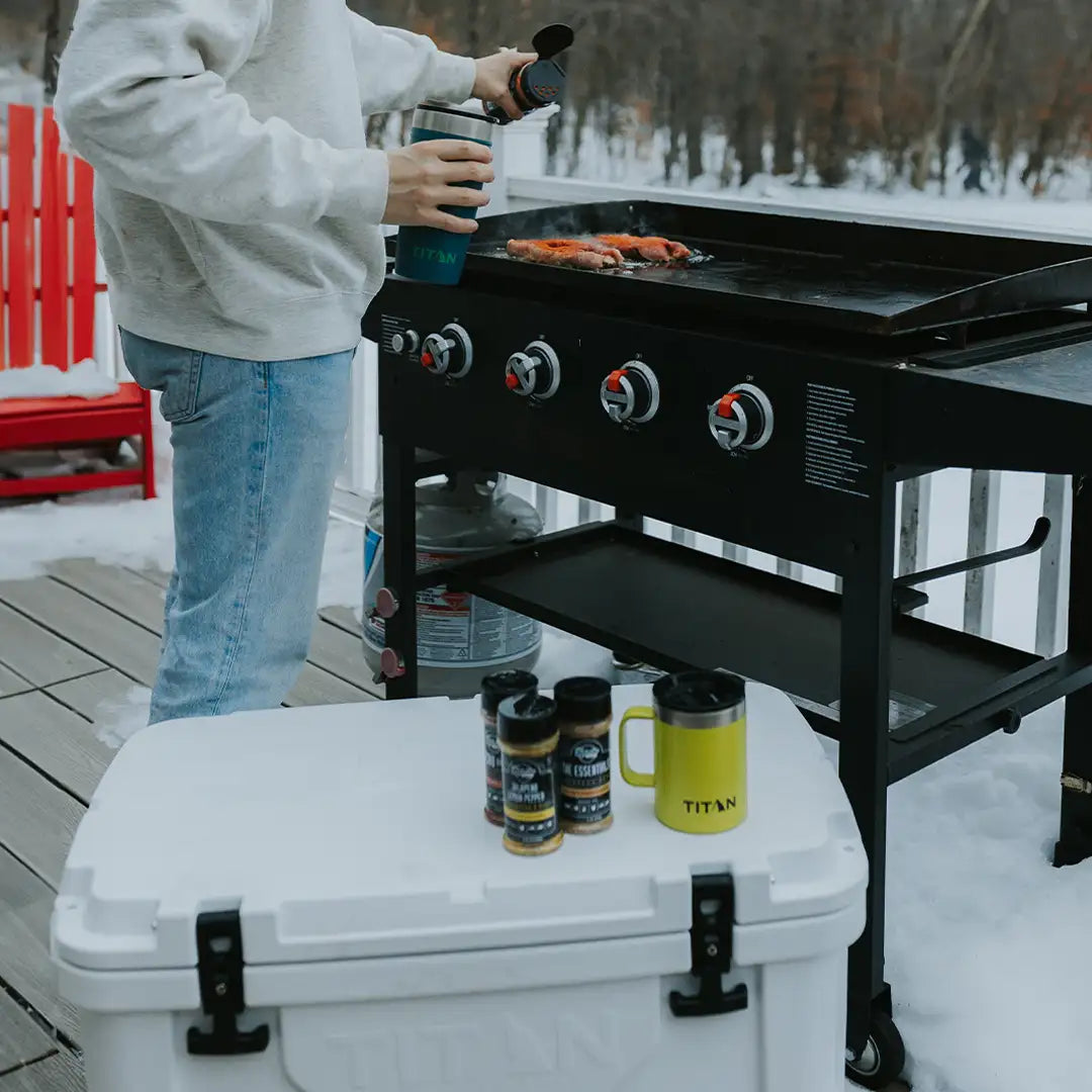 Person grilling food on a large outdoor grill beside a white hard cooler.