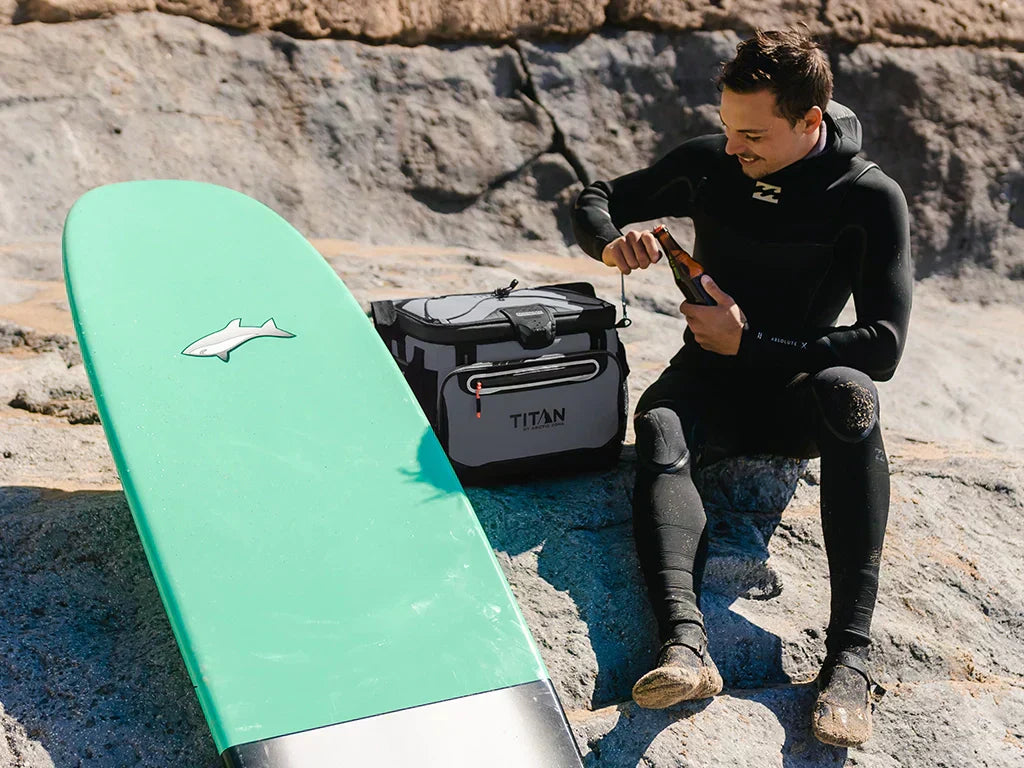 A guy enjoying a cold drink next to a Titan Cooler and surfboard at the beach