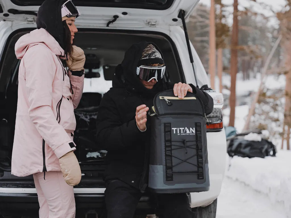 Two people grabbing a cold drink from a Titan Welded Backpack Cooler after skiing