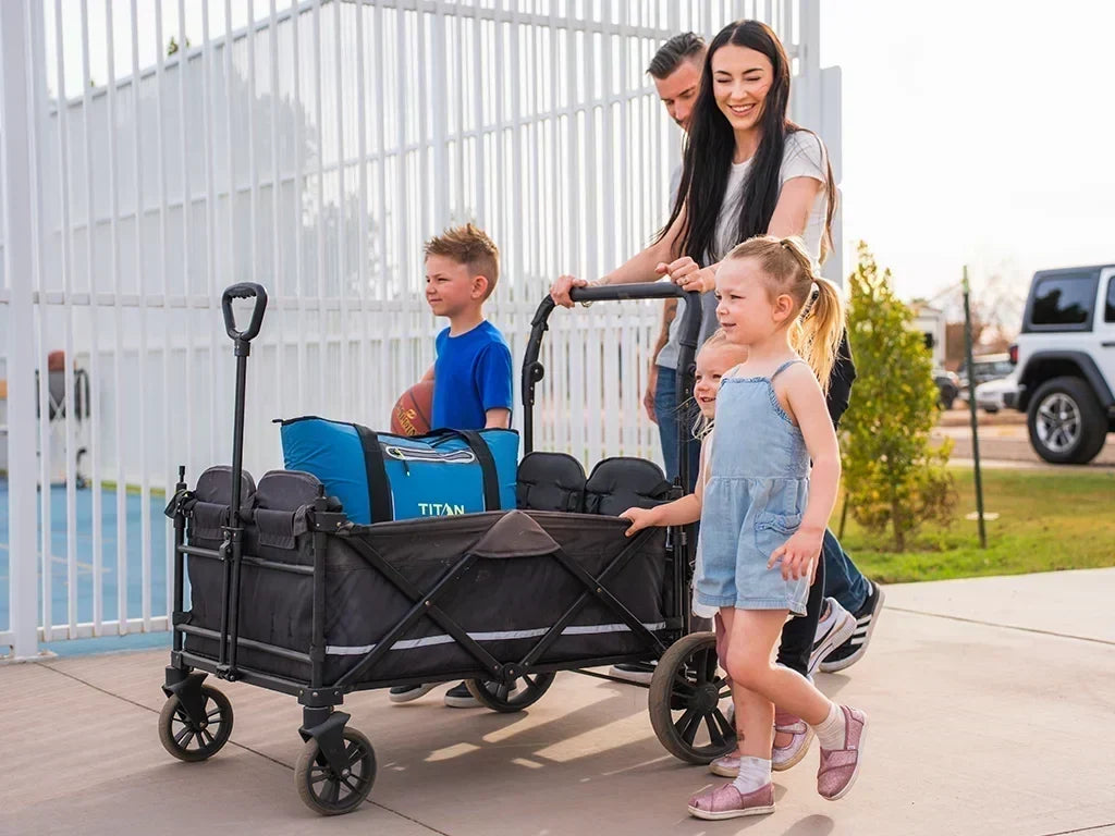 Two adults and two children walking beside a black folding wagon with an insulated tote inside along a paved walkway.