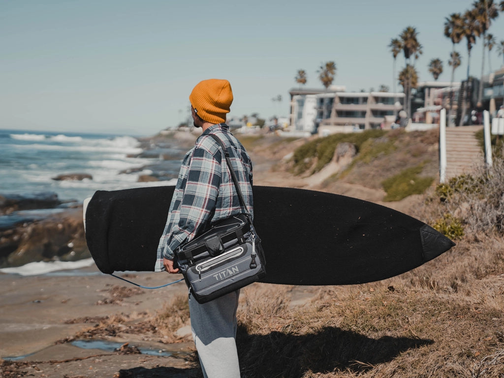 Guy at the beach with a surf board and Titan 40 Can Collapsible Cooler