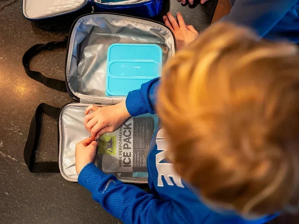 Top-down view of an open insulated lunch bag with a blue food container and reusable ice pack while a child packs the bag.
