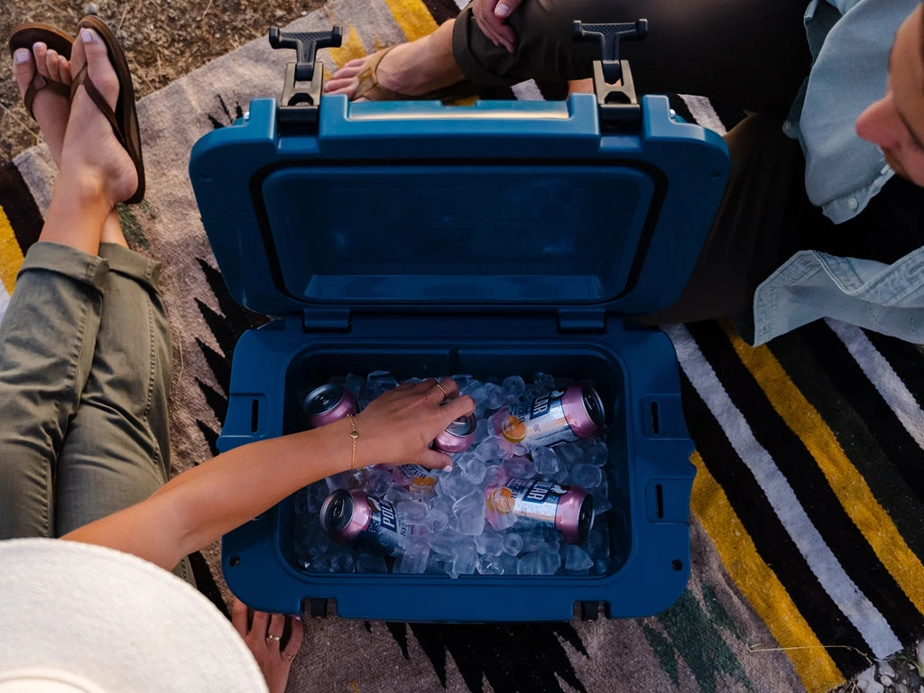 Two people sitting on a picnic blanket with a hard cooler full of cold drinks and ice