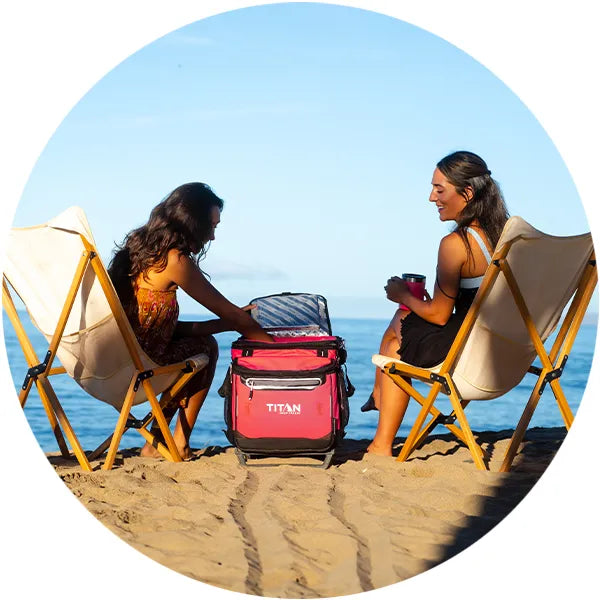 Two women sitting at the beach with a Titan Wheeled Cooler 