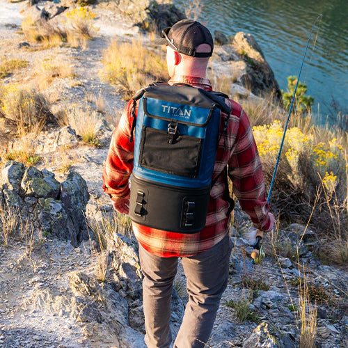 A man with a fishing rod, wearing a Titan Backpack Cooler by a lake