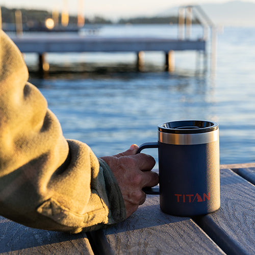 A man holding a Titan mug while sitting on a dock looking out at the lake