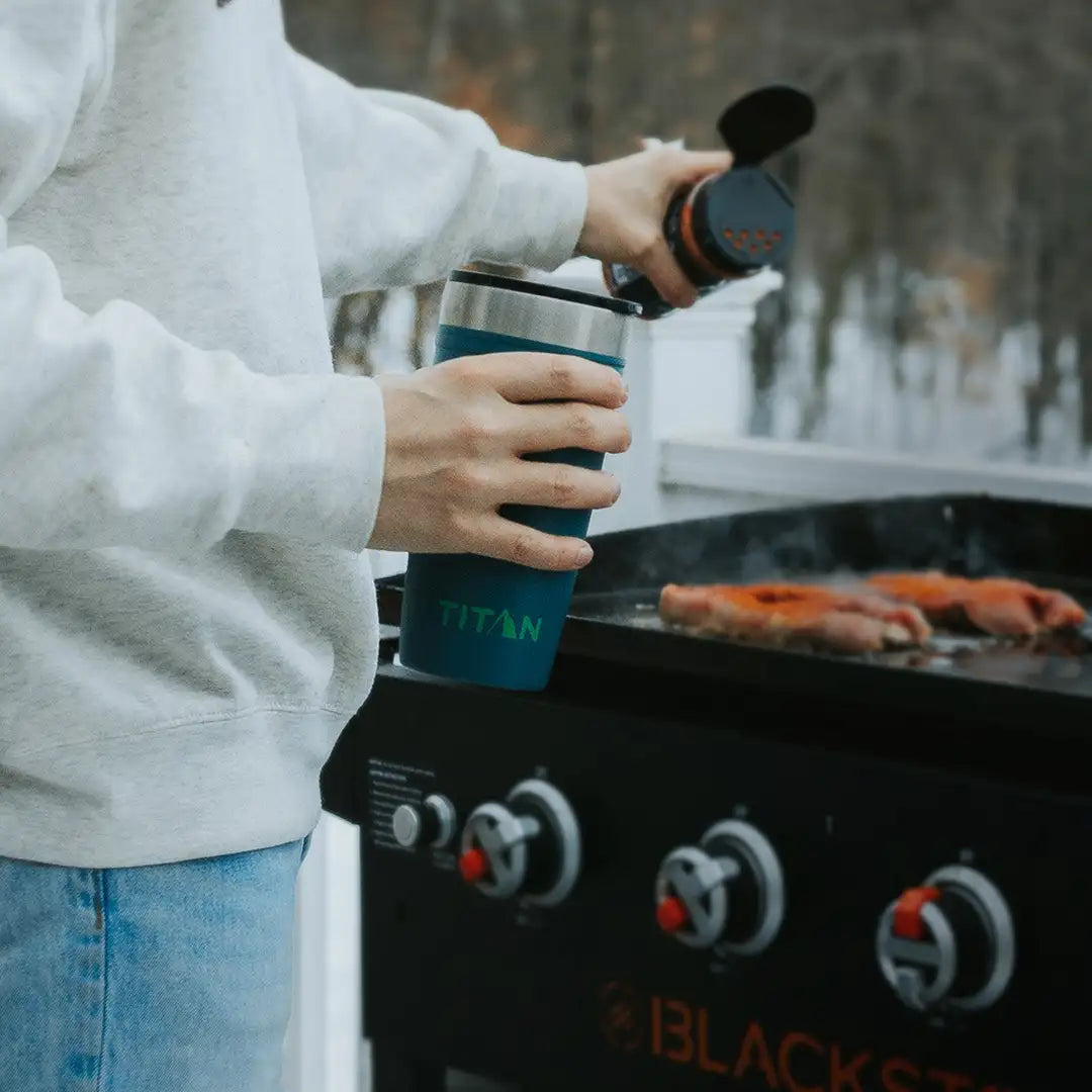 Person holding a tumbler in hand while seasoning food on a grill