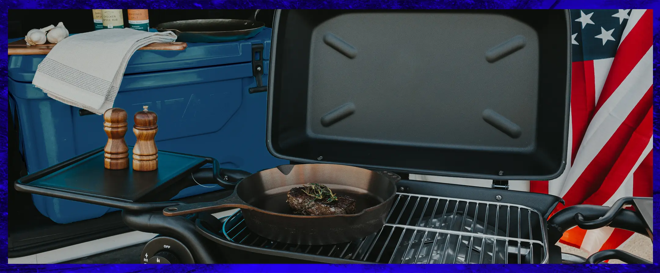 A portable grill with a steak in a skillet, next to a hard cooler and American flag.